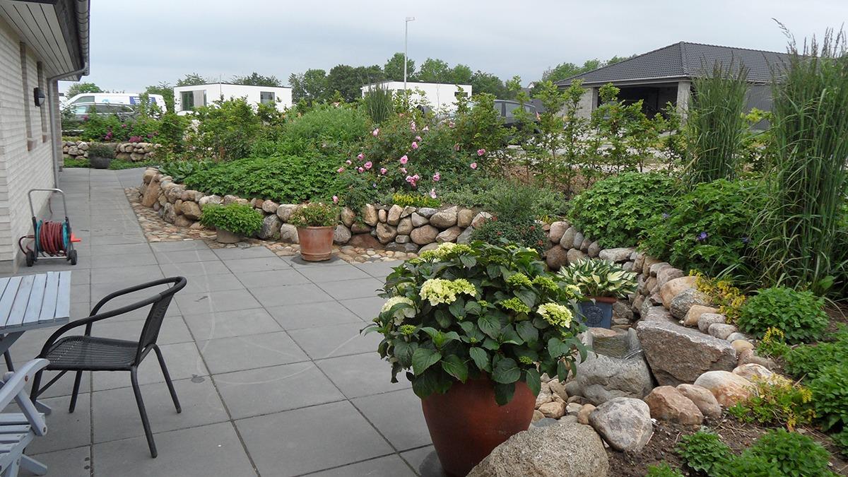 En smuk have med blomster, buske og en stenmur, set fra en terrasse med havemøbler.