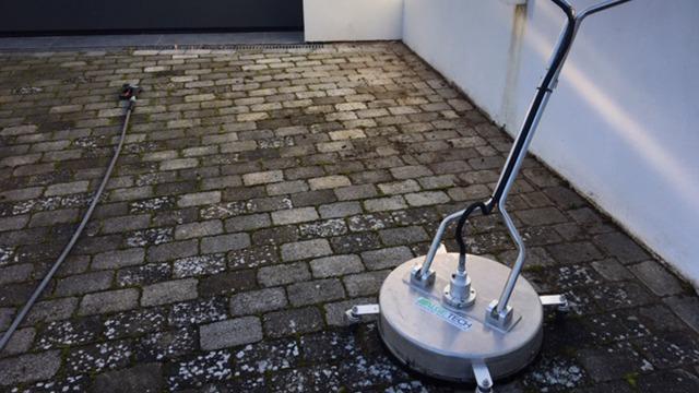 En højtryksrenser med en rund børste står på en flisebelagt overflade ved en væg.