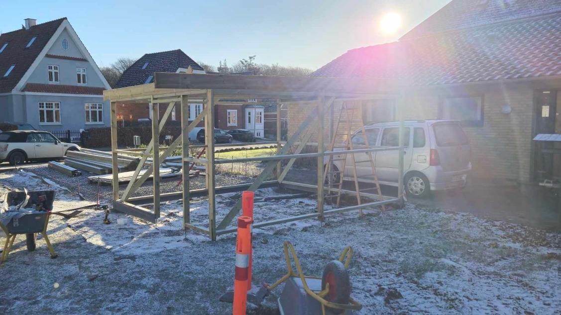 Byggeplads med træramme til carport, bil parkeret, solskin og sne på græsset.