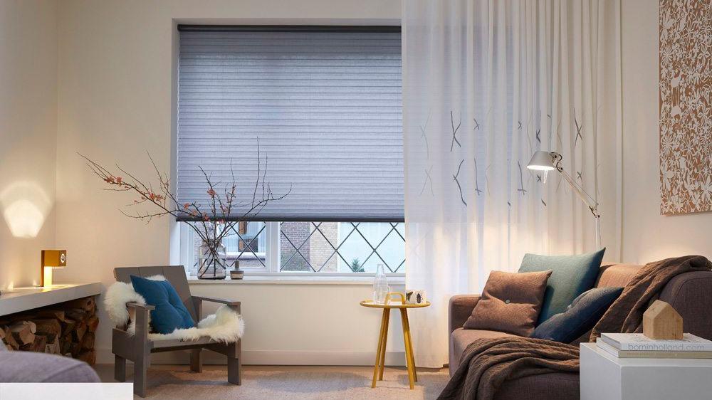 Moderne stue med grå persienner, bløde puder, en lænestol og en lille sidebord ved vinduet.