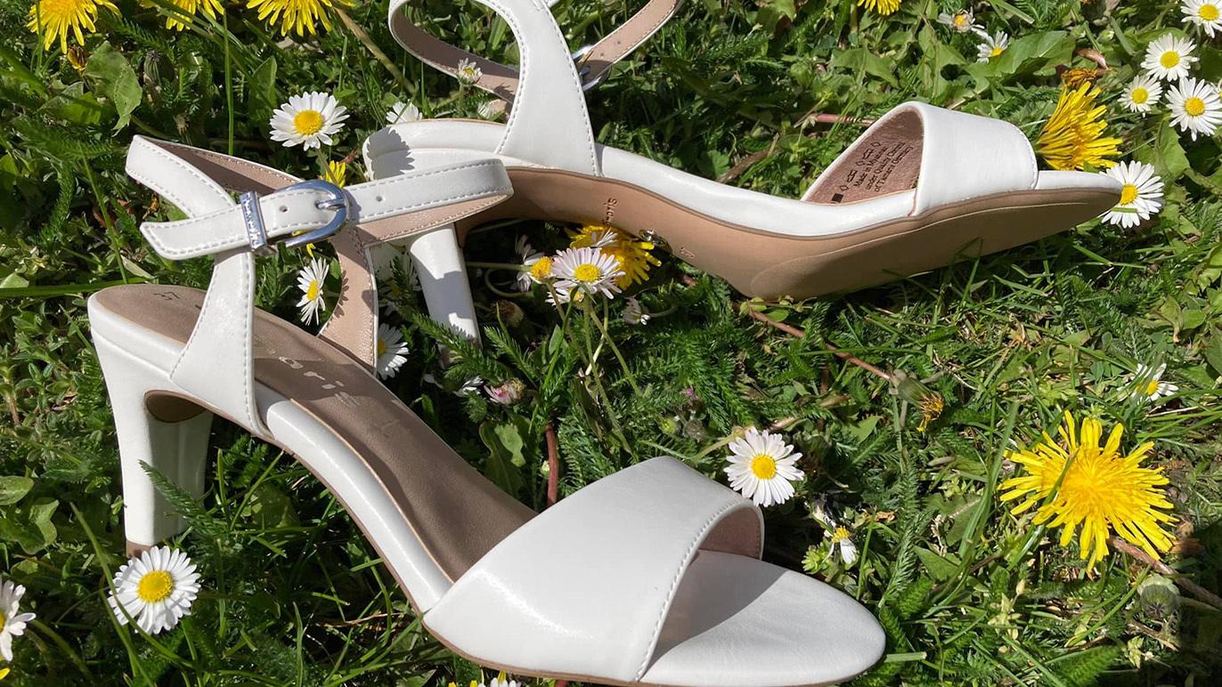 Hvide højhælede sandaler placeret på græsset blandt blomster.