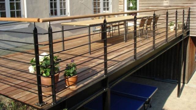 Terrasse med trædæk, metalgelænder og borde med stole, omgivet af blomsterpotter.