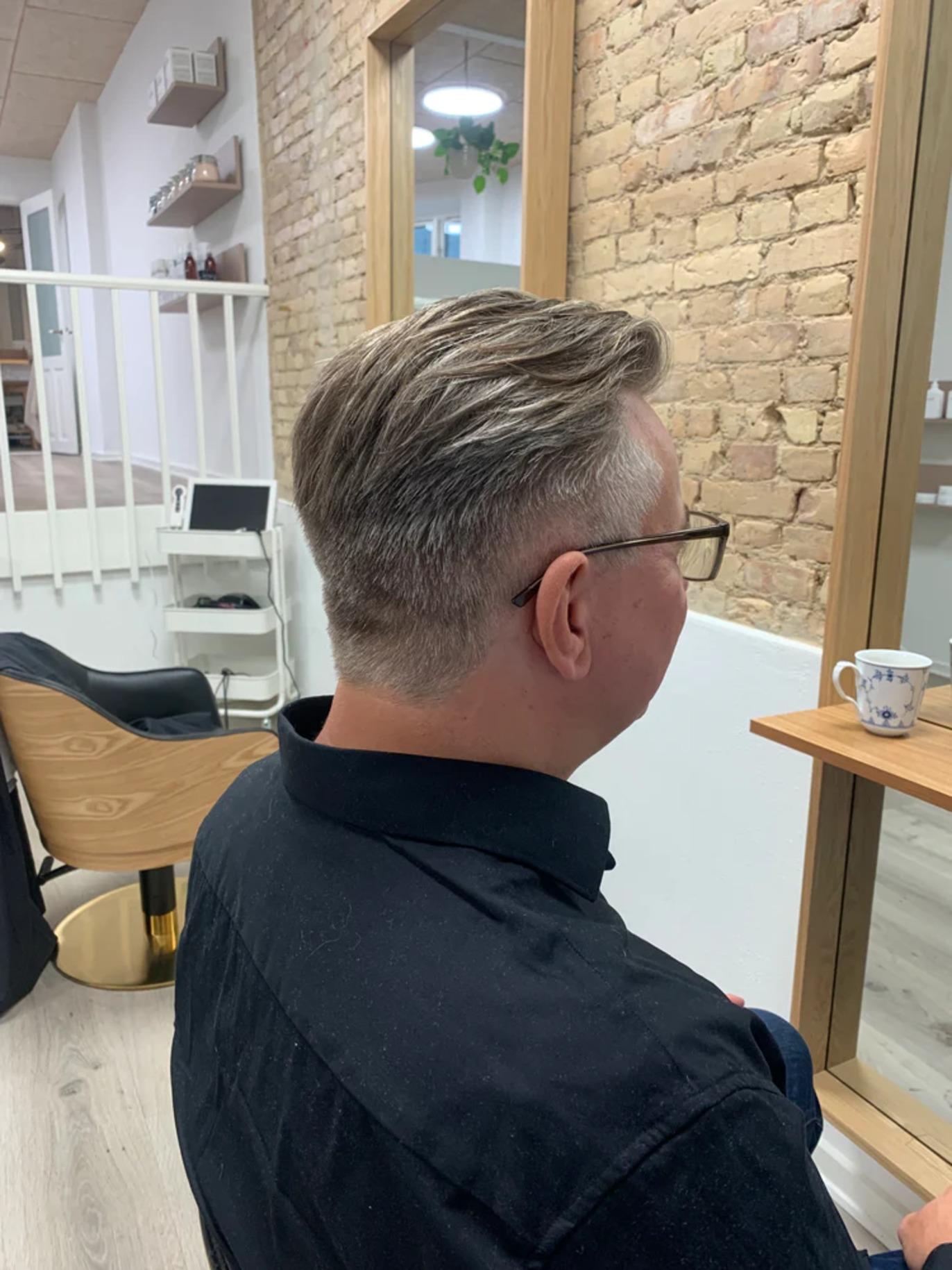 Mand med kort hår og briller, der sidder i en frisørsalon med murstensvæg.