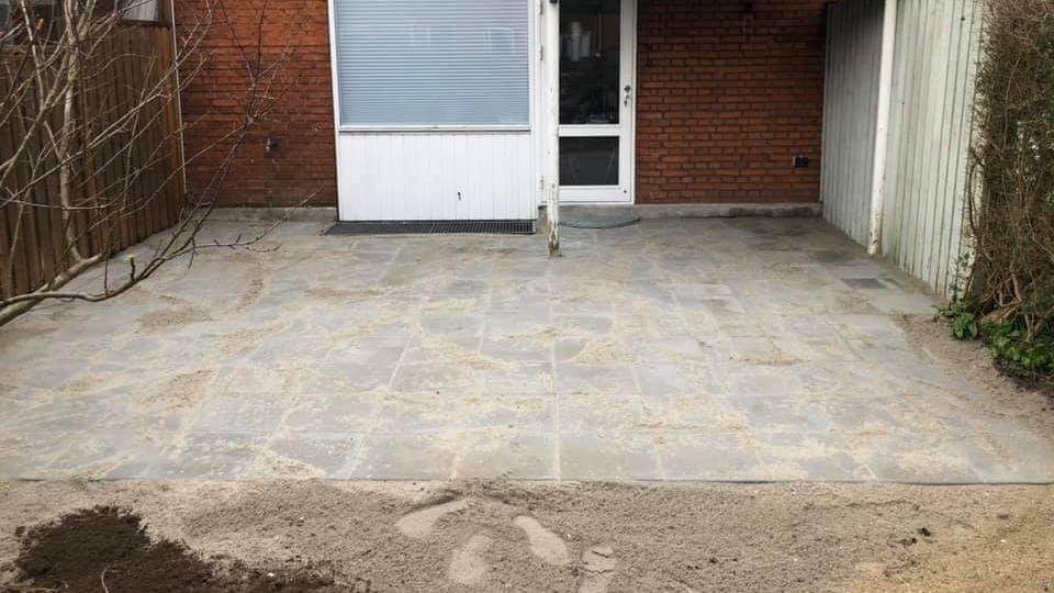 Nybelagt flisebelægning foran en husfacade med en dør og vindue. Sand og jord i forgrunden.