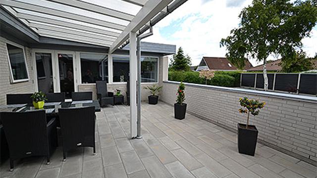 En moderne terrasse med glasoverdækning, havemøbler og grønne planter i krukker.