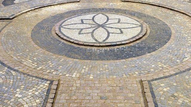 Mosaikbelægning med cirkulære mønstre og en blomst i midten, lavet af forskellige sten i nuancer af grå og beige.