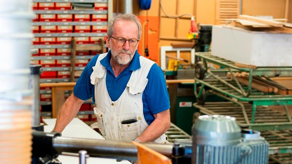 En ældre mand i arbejdsdragt arbejder koncentreret i et værksted med værktøj og materialer omkring sig.