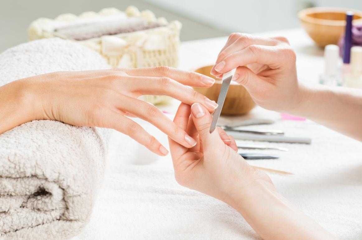 Håndpleje med neglefilning på en manicurebord med håndklæde og manicureværktøjer i baggrunden.