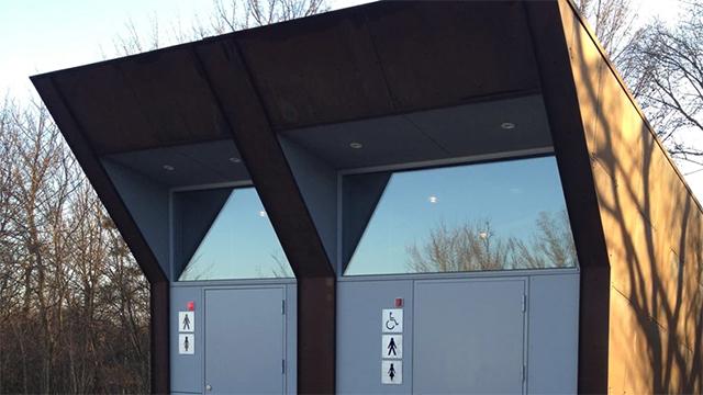 Moderne toiletbygning med to indgange, handicapvenlig adgang og store vinduer i et skovområde.