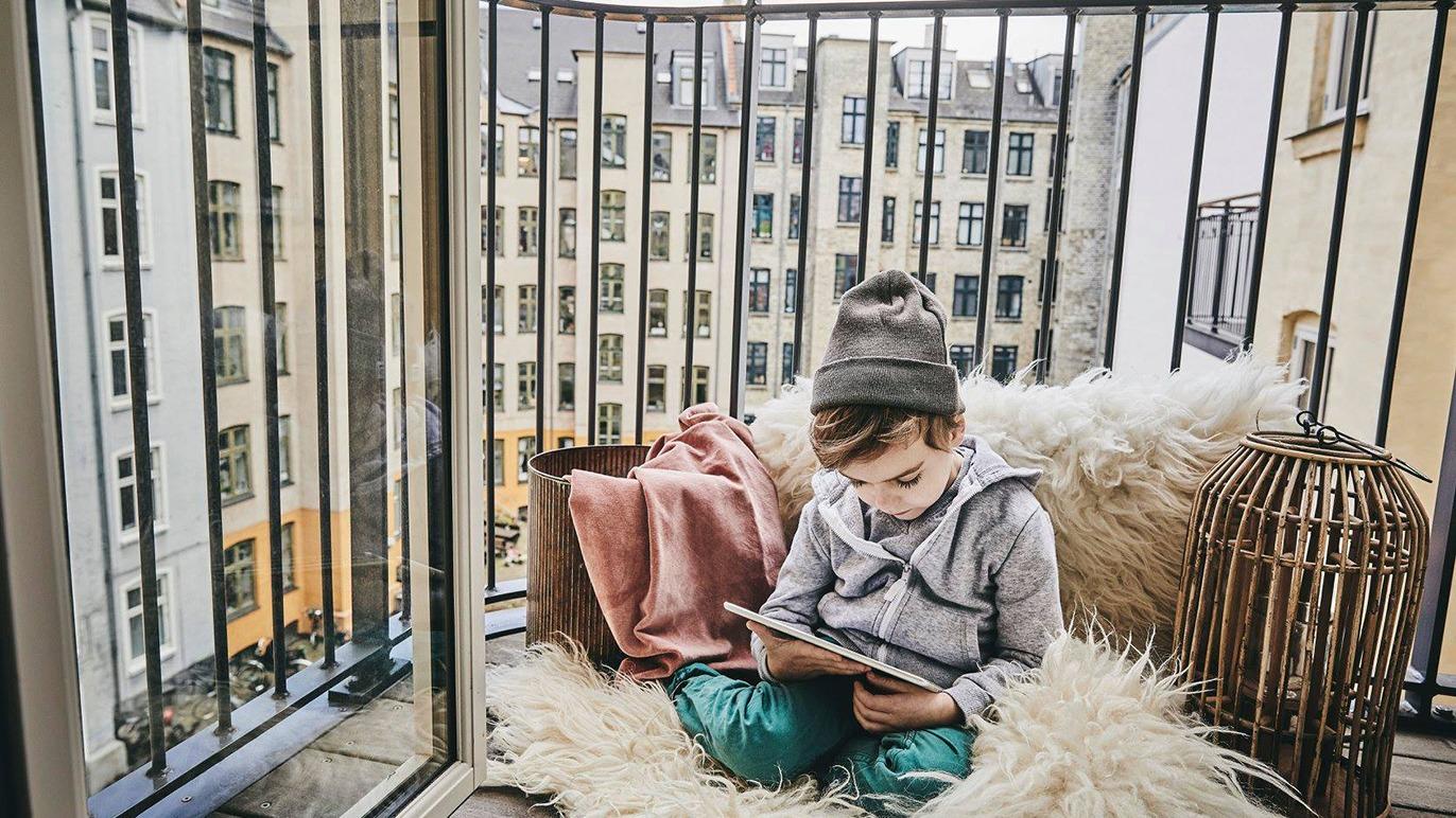 Barn sidder på en balkon i en hyggelig krog med en tablet, omgivet af puder og tæpper.