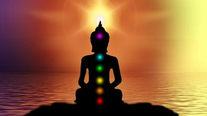 En silhuet af en Buddha, der mediterer ved vandet med farverige chakraer og en solopgang i baggrunden.