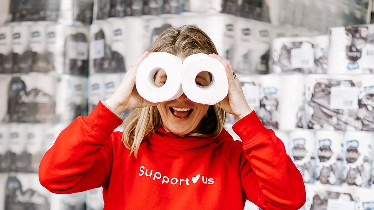 Kvinde iført rød sweatshirt holder toiletpapir foran ansigtet som briller, smilende i en lagerhal.