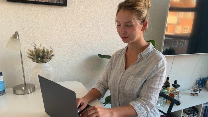 En ung kvinde sidder ved et skrivebord og arbejder på en bærbar computer i et lyst rum.