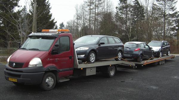 En lastbil med tre biler på en trailer, parkeret på en grusvej omgivet af træer.