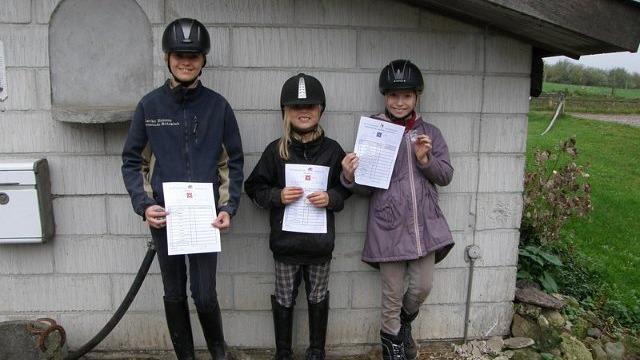 Tre børn i rideudstyr står foran en bygning og holder deres certifikater.