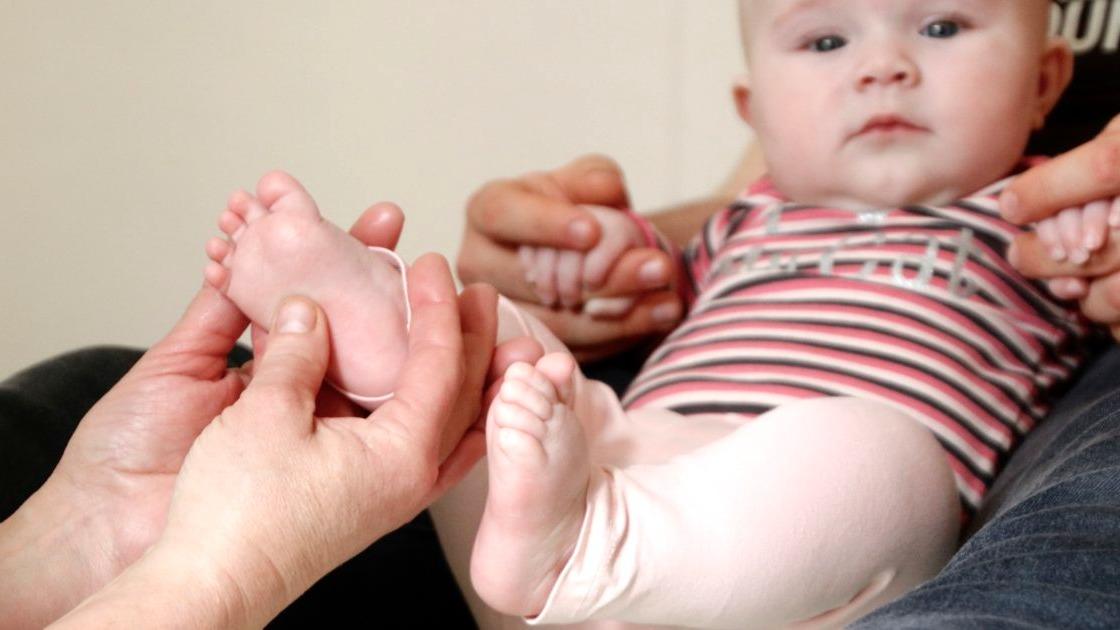 En baby med stribede tøj sidder på skødet af en voksen, mens en anden voksen holder dens fødder.