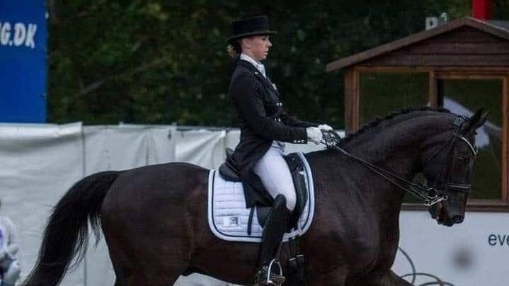 En rytter i sort dressuroutfit rider på en sort hest under en konkurrence i en udendørs arena.