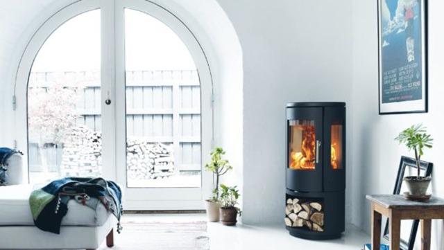 Moderne brændeovn i stue med hvid væg, sofa og store vinduer, der giver lys ind i rummet.