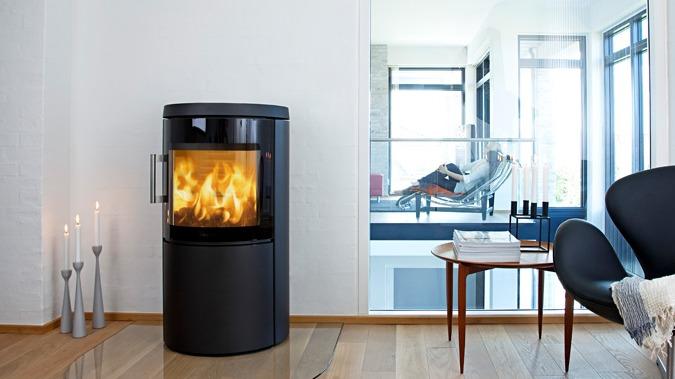Moderne brændeovn i stue med flammer, lysestager og et stilfuldt møbel i baggrunden.
