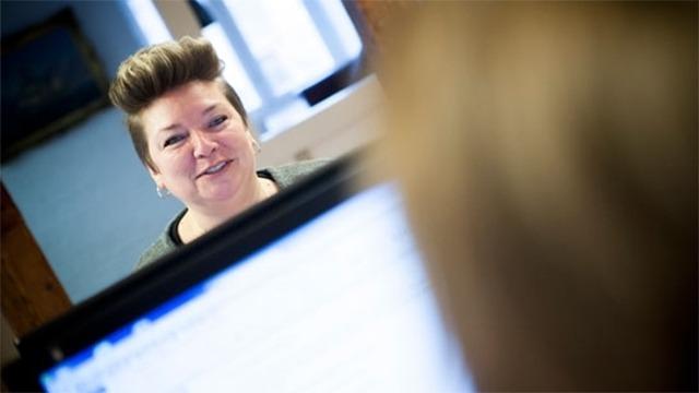 En kvinde med kort hår smiler, mens hun taler med en person bag en computer.