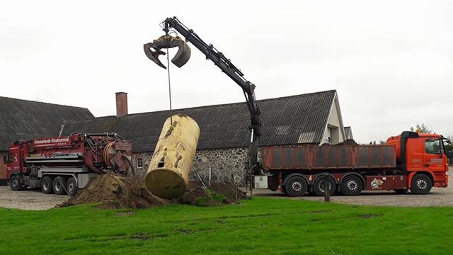 En kran løfter en stor gul tank fra jorden ved en gård, mens to lastbiler står parkeret i baggrunden.