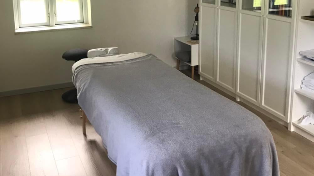 Et massageværelse med en massagebriks dækket af et gråt tæppe og et vindue, der giver naturligt lys.