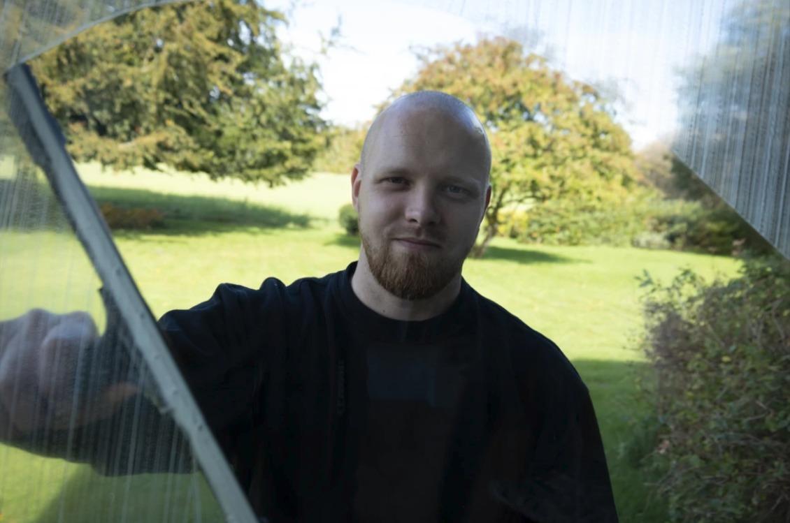 Mand med skaldet hoved og skæg står ved vinduet og kigger ud på en grøn have med træer.