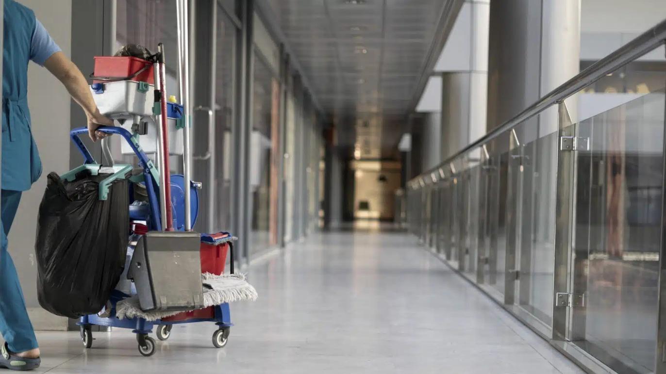 En sygeplejerske skubber en rengøringsvogn ned ad en moderne hospitalsgang med glasvægge.
