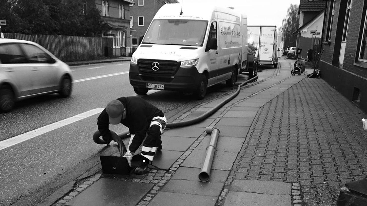 En mand arbejder på fortovet med en bærbar computer, mens en varevogn holder parkeret i baggrunden.