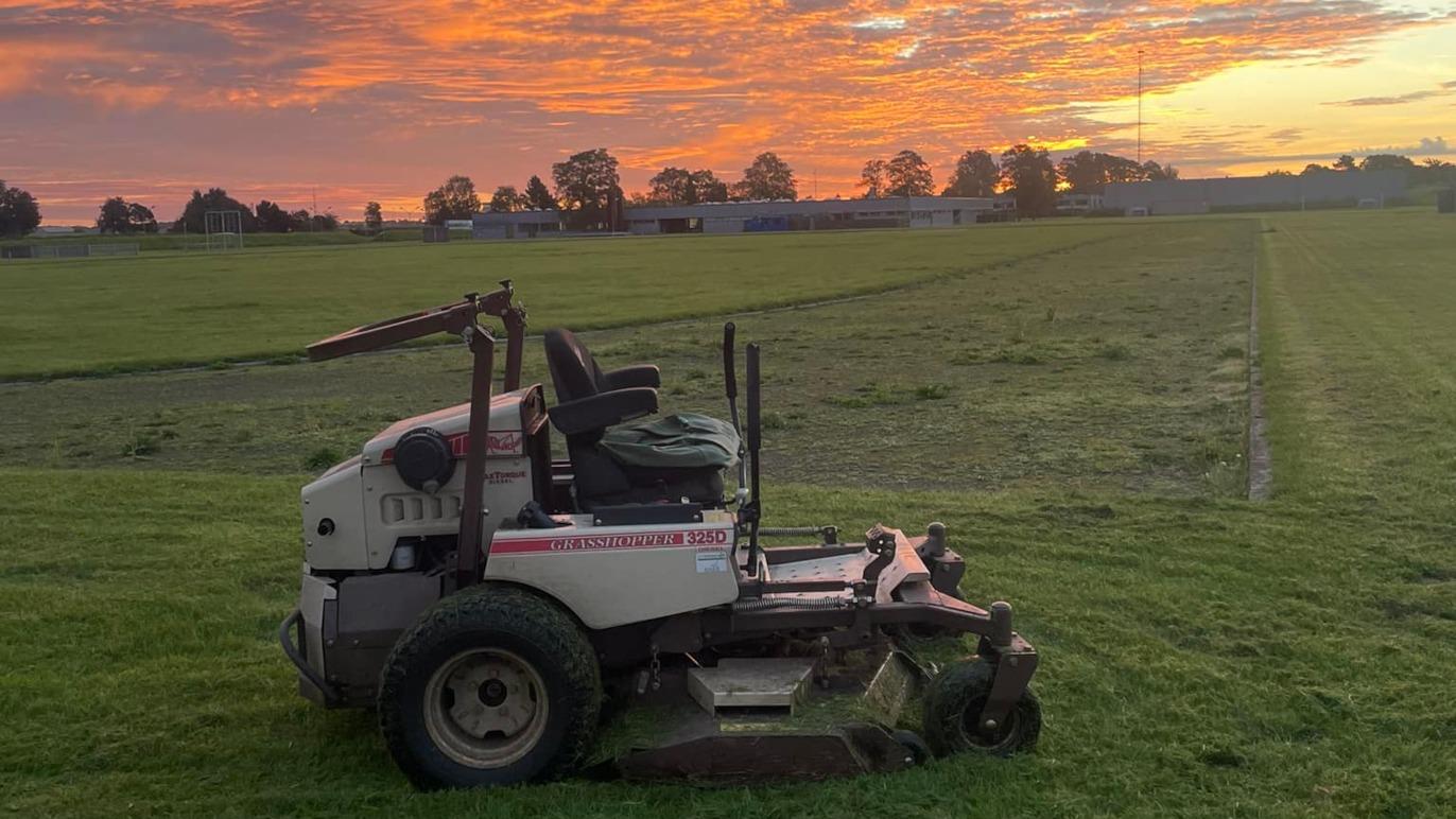 En græsslåmaskine står på en nyklippet græsmark med en farverig solnedgang i baggrunden.