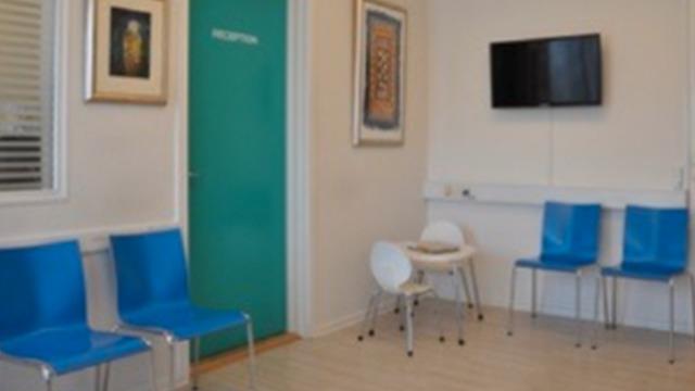 Modtagelsesområde med blå stole, et lille bord og en vægmonteret tv-skærm. Døren til receptionen er grøn.