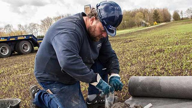 En mand i hjelm arbejder på en mark, mens han undersøger jorden med en pose og værktøj.