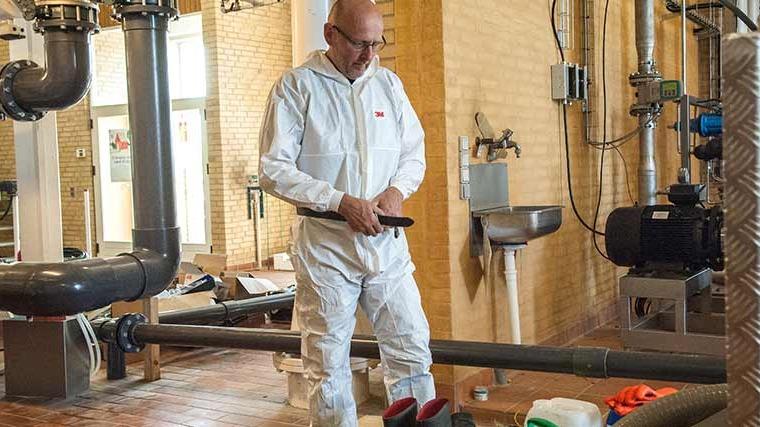 En mand iført en hvid beskyttelsesdragt arbejder i et industrielt miljø med rør og udstyr.