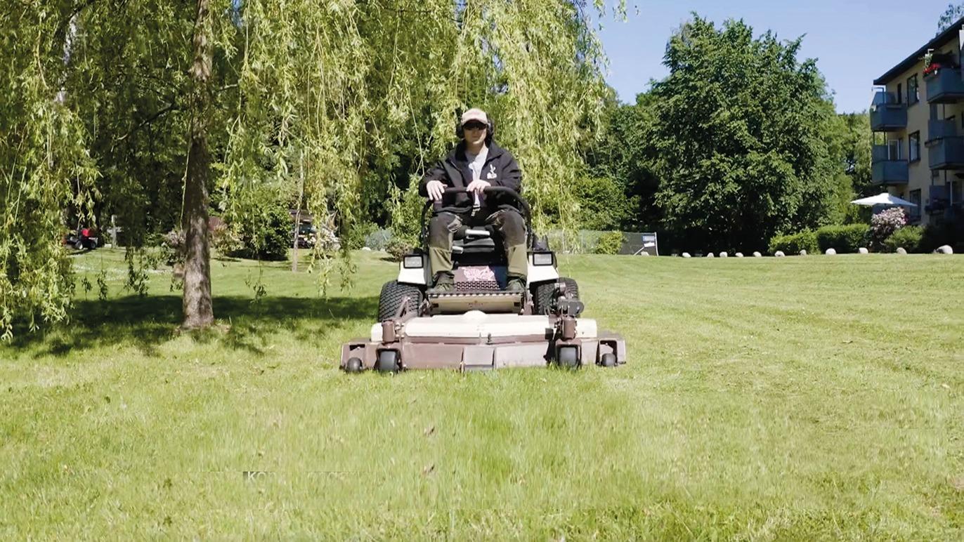 En person klipper græs med en rideklipper på en grøn græsplæne under en solrig dag.