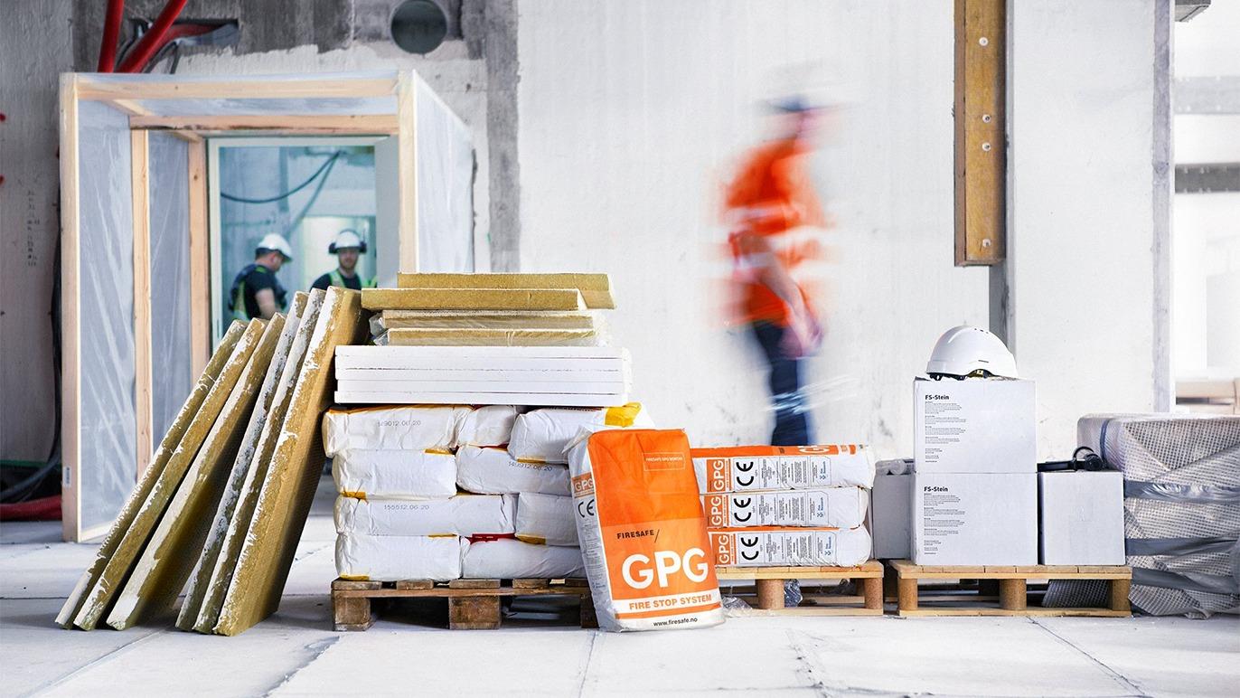 Byggeplads med materialer som gipsplader, isolering og cement, mens håndværkere arbejder i baggrunden.