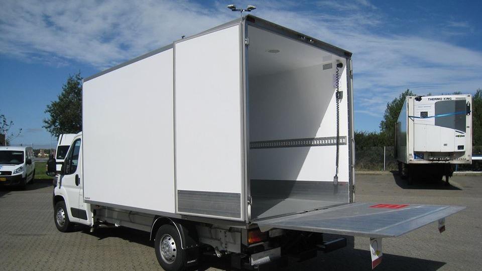 En hvid lastbil med åben bagklap og tom lad, parkeret på en gårdsplads under en blå himmel.