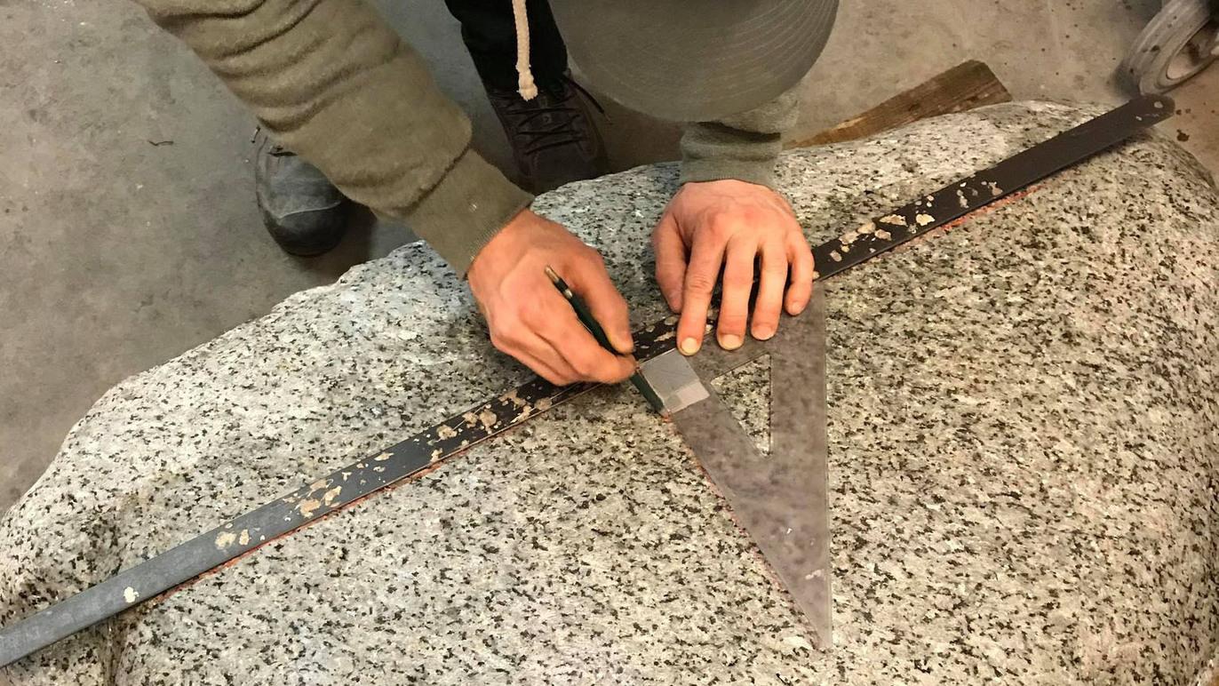 Hænder der bruger en vinkelmåler til at måle på en granitplade i et værksted.
