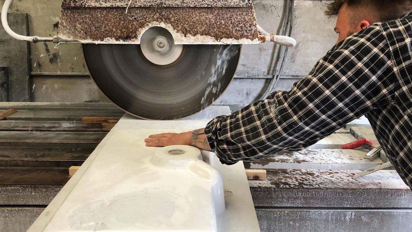 En håndværker skærer en stor marmorplade med en sav i et værksted. Støv og materialer flyver omkring.