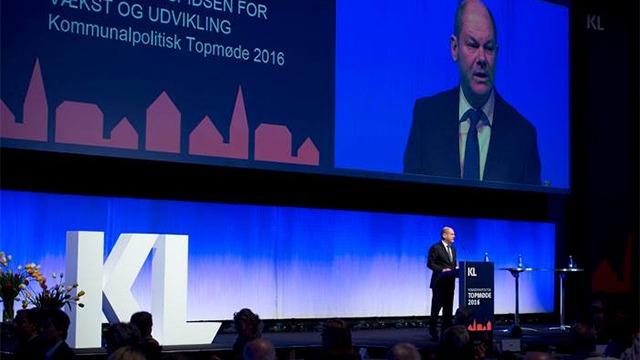En mand taler ved en konference med en stor skærm i baggrunden, der viser teksten "KL Kommunalpolitisk Topmøde 2018".
