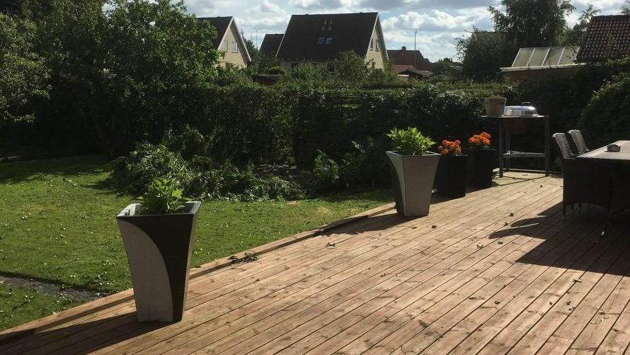 En solrig terrasse med trædæk, blomsterkrukker og en velplejet have i baggrunden.