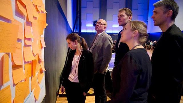 Fem personer ser på orange post-it sedler på en væg under en workshop.