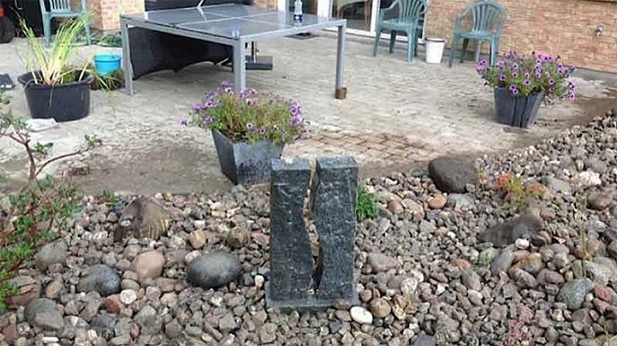 En have med en stenfigur, blomsterkrukker og en terrasse med havemøbler.