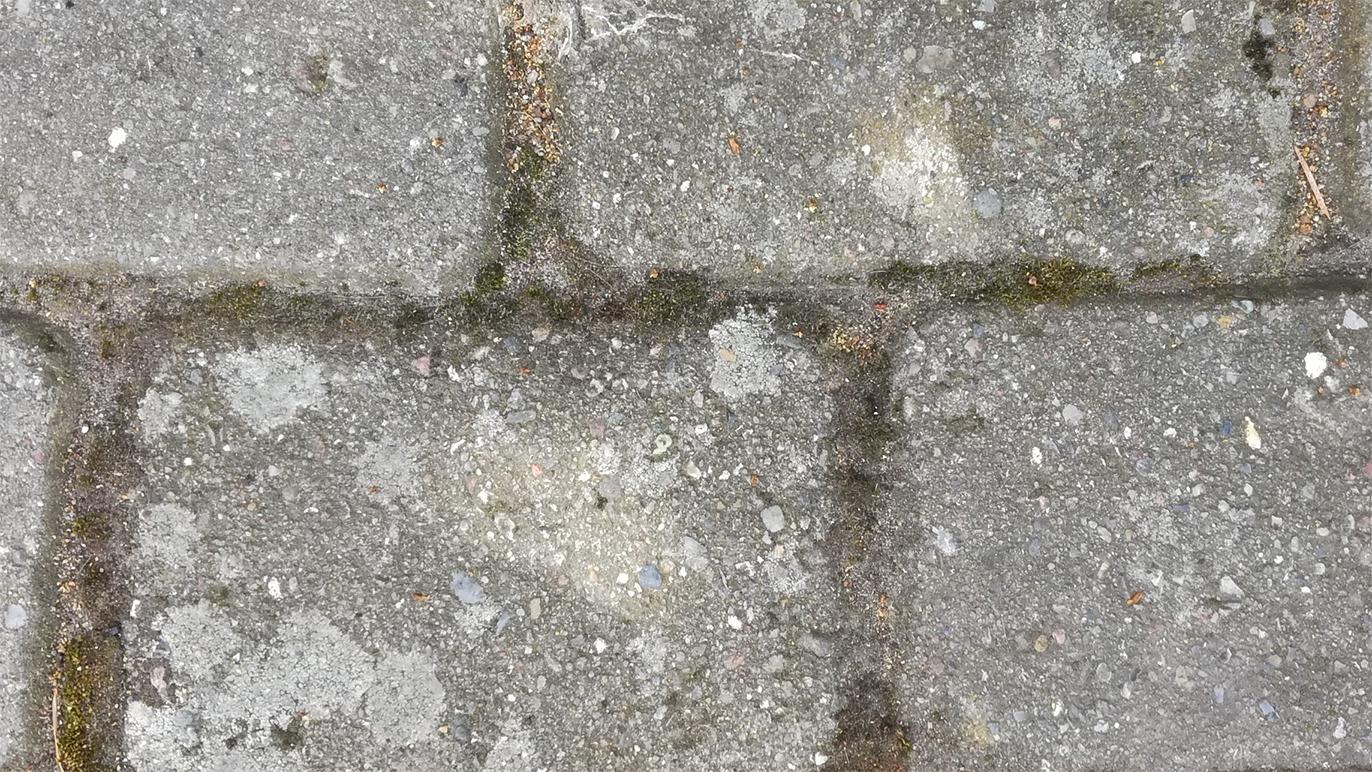 Gammel brostenbelægning med græs og småsten imellem, der viser slid og naturlig patina.
