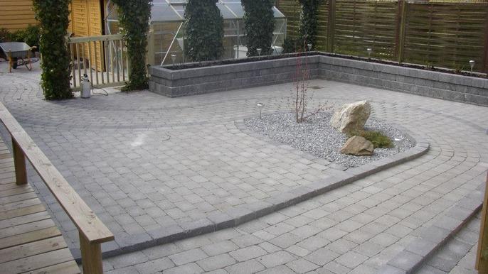 En velanlagt terrasse med grå fliser, småsten og en dekorativ sten i midten, omgivet af grønne planter.