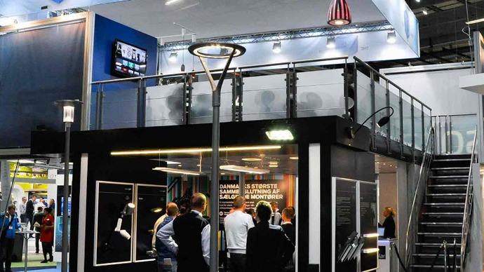 En moderne messestand med glasvægge og en trappe, hvor besøgende interagerer med udstillere.