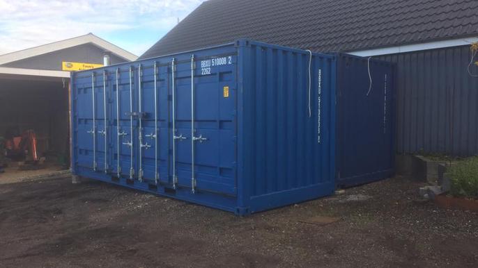 To store containerne i en gård med en bygning i baggrunden og grus under.