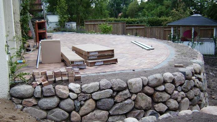 Terrasse med stenbelægning og en stensokkel, der er under konstruktion i en have. Kasser og materialer er synlige.
