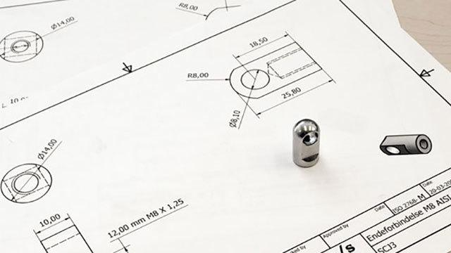 Teknisk tegning med en metalkomponent placeret på papiret, der viser mål og detaljer.