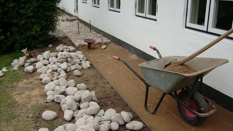 En skovl med sand og en skubbevogn ved siden af en sti med store sten langs en hvid væg.