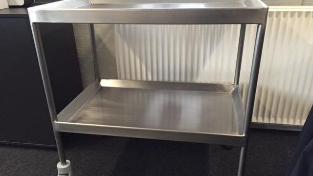 Et stålvogn med to hylder, placeret på hjul, står foran en radiator.